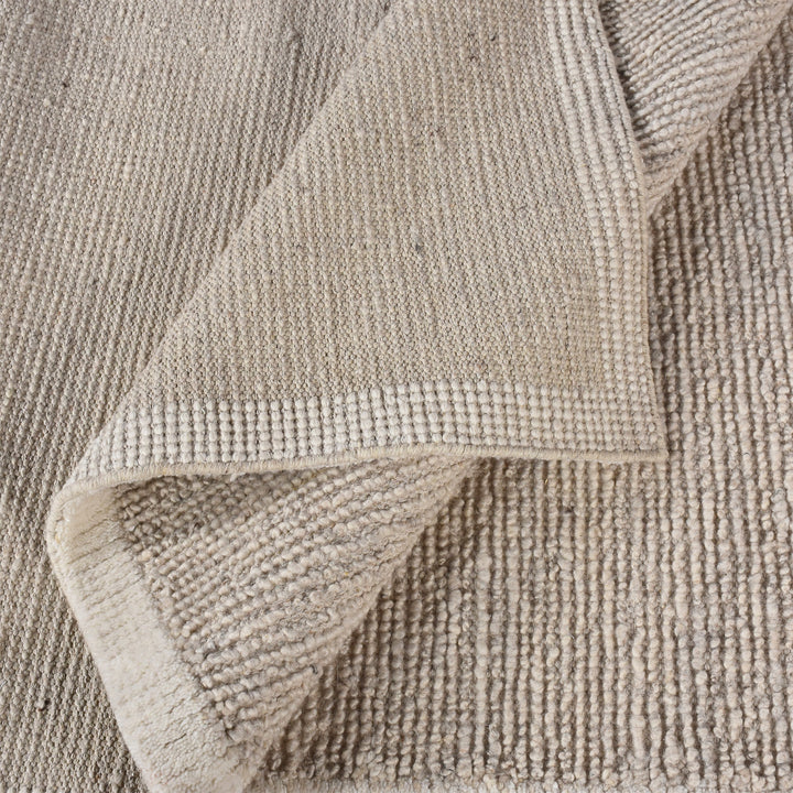 Shant handwoven flatweave wool rug in natural ivory with fine ribs and fringe, beside a bed